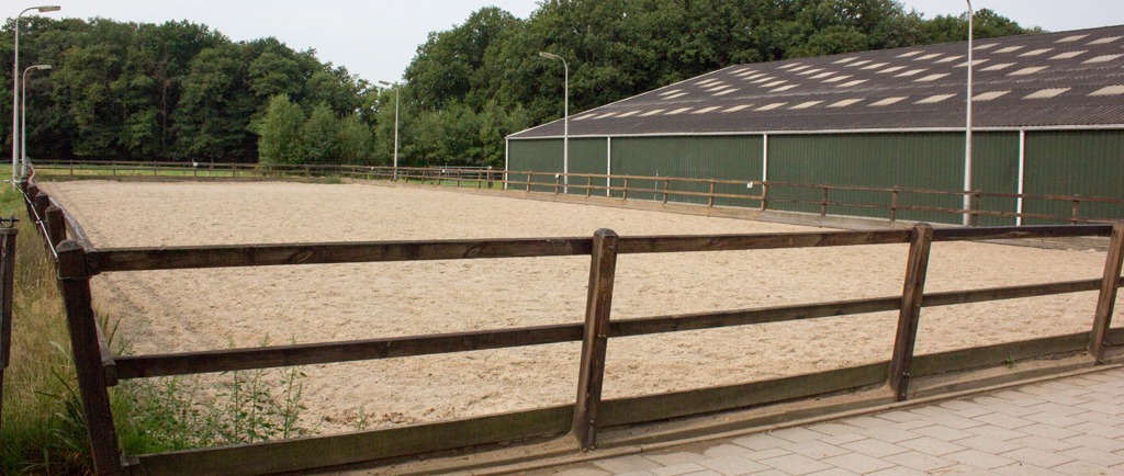 De all-weather buitenbak met zandbodem en houten omheining, naast de groene binnenmanege.